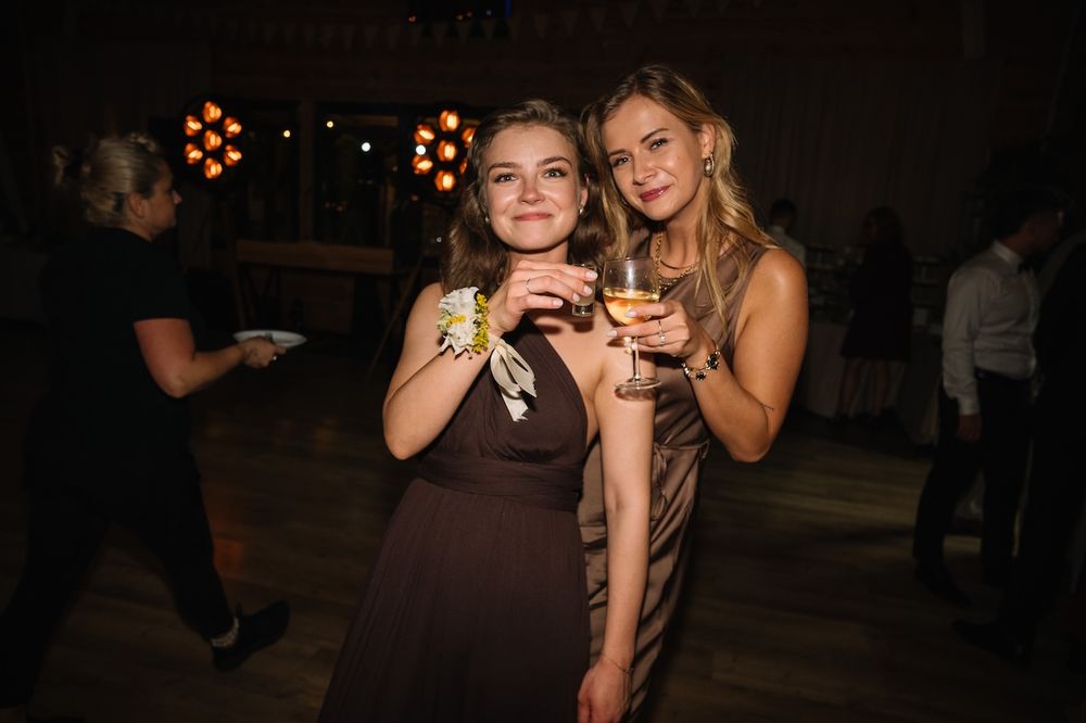 Two women in brown dresses smile holding wine glasses at a wedding reception.