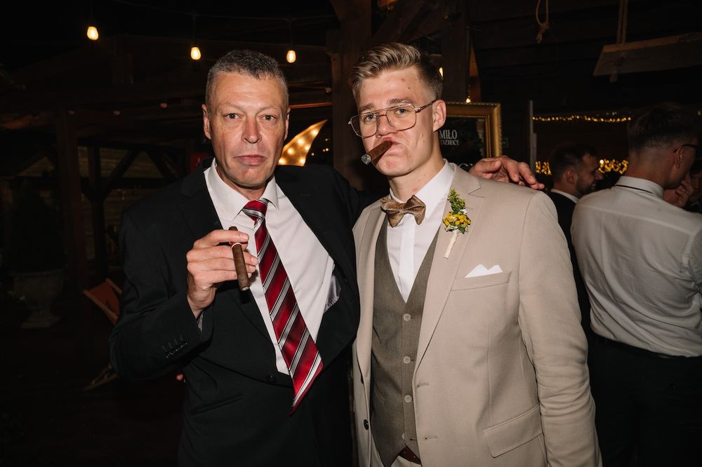 Two men pose with cigars at a wedding reception, one wearing a bow tie.