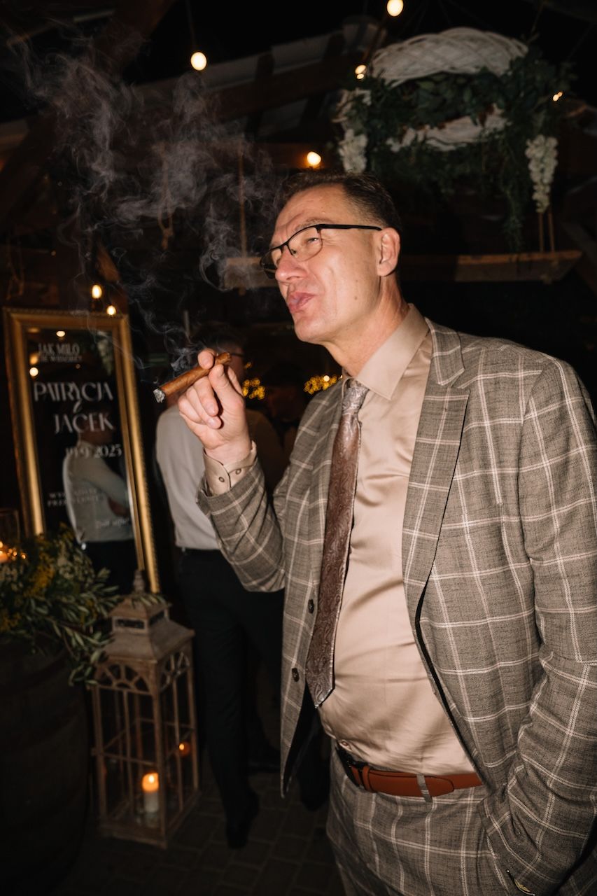 Elegant man in plaid suit smoking a cigar at an indoor wedding reception.