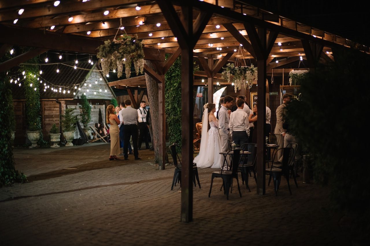 Wedding guests mingle under a wooden pergola with string lights at night.