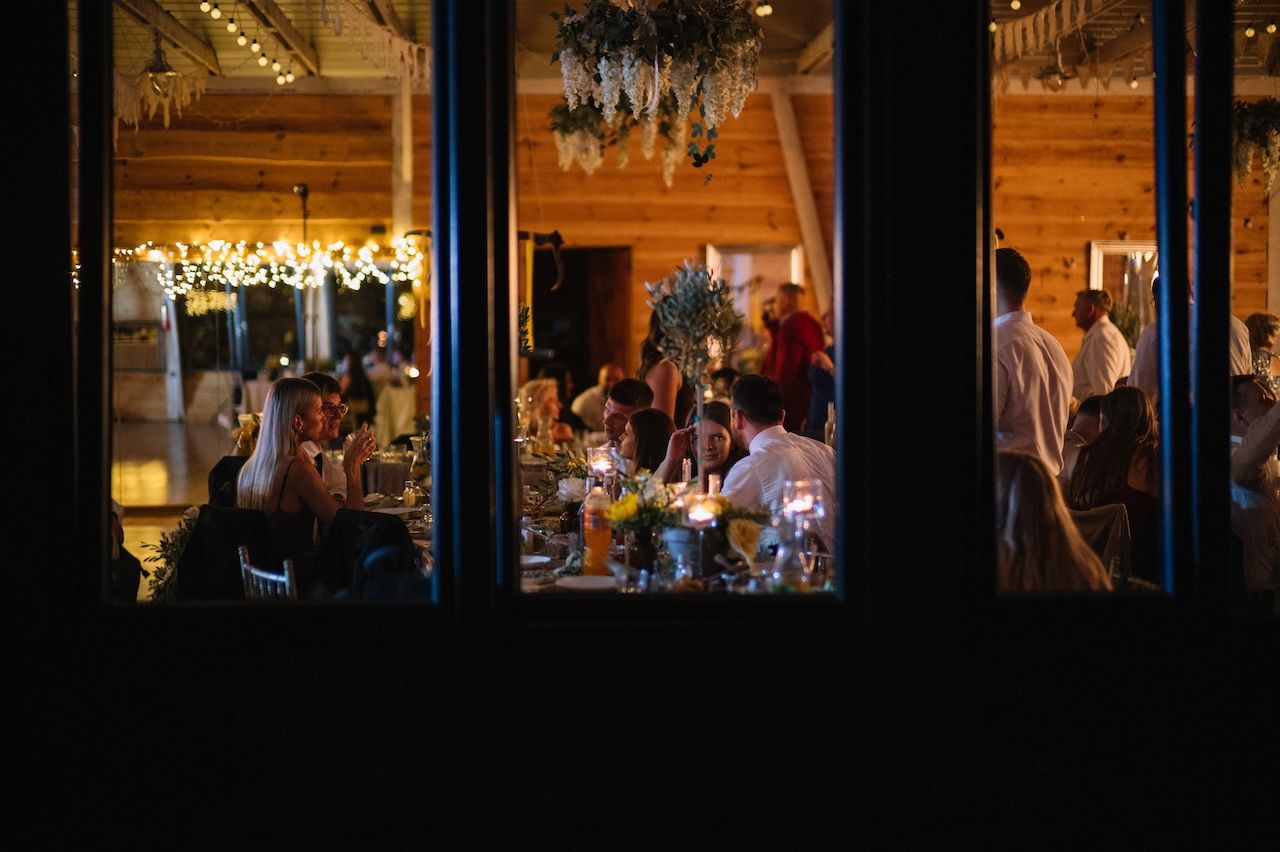 Wedding reception dinner viewed through large windows with guests seated at candlelit tables.