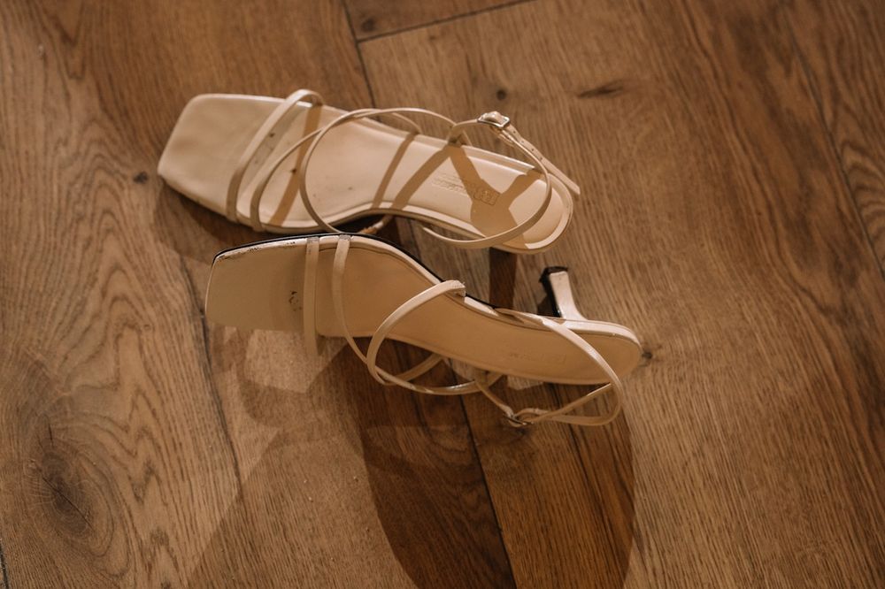 Pair of beige strappy heeled sandals resting on a wooden floor at a wedding.