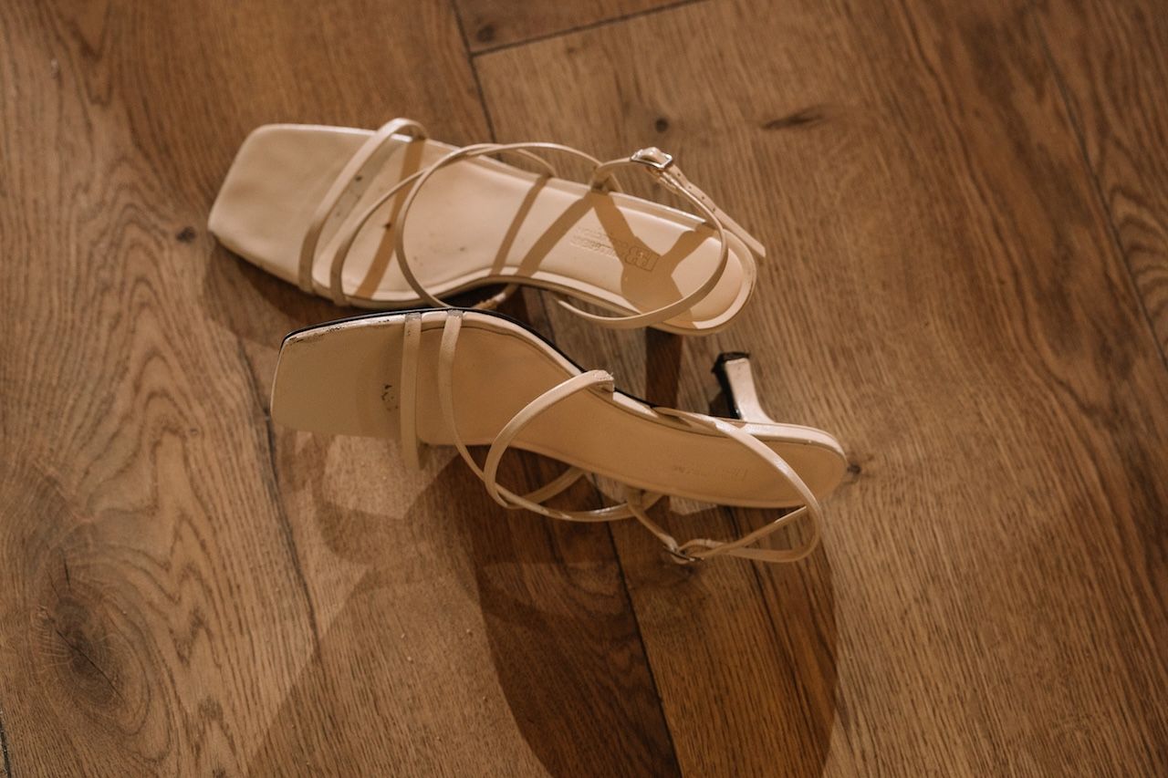 Pair of beige strappy heeled sandals resting on a wooden floor at a wedding.