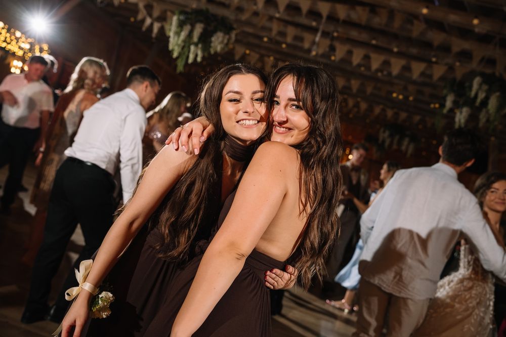 Two young women in brown bridesmaid dresses hugging and smiling at a wedding reception.