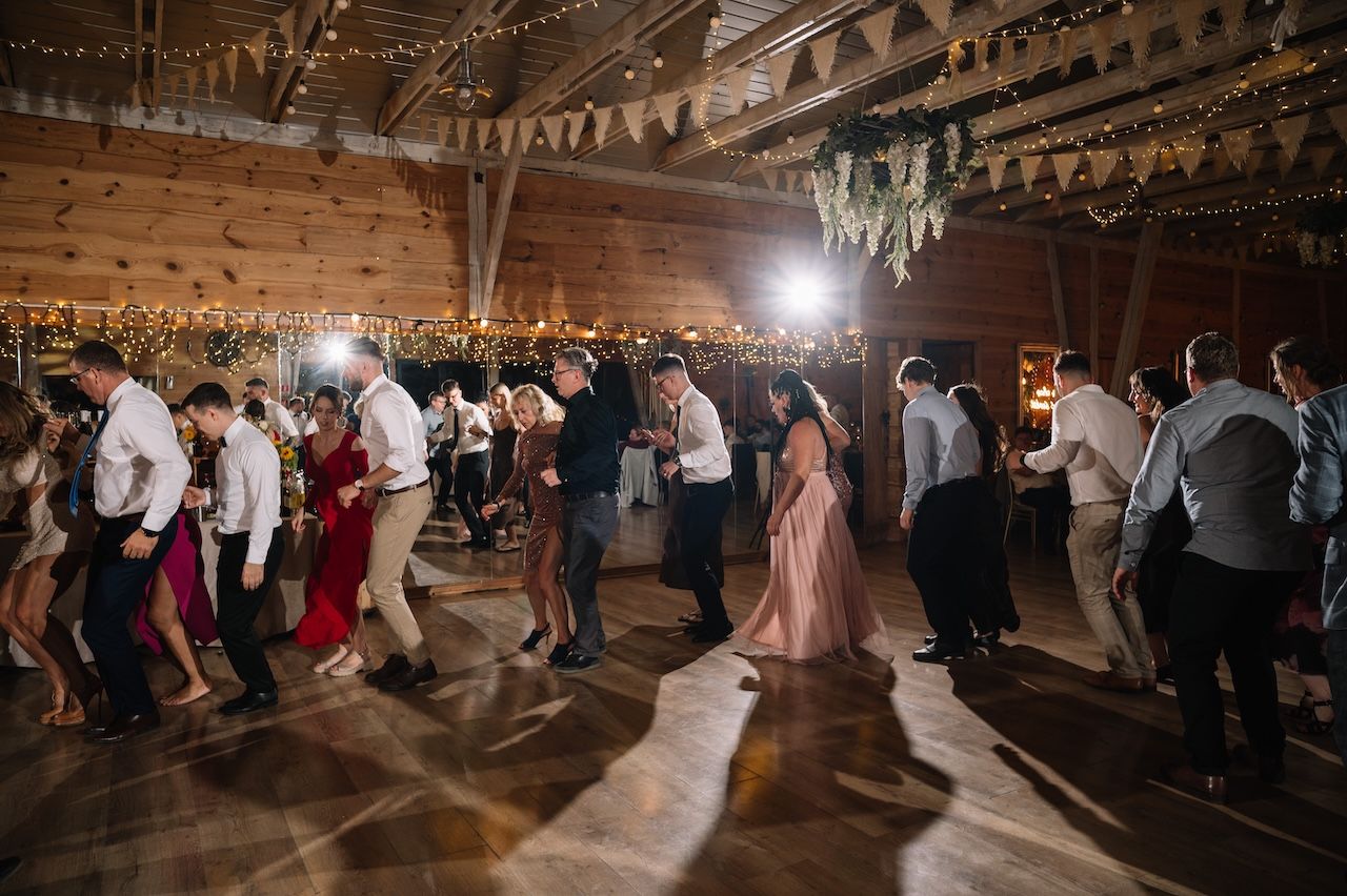 Large group of wedding guests dancing on a wooden floor in a barn venue with string lights.