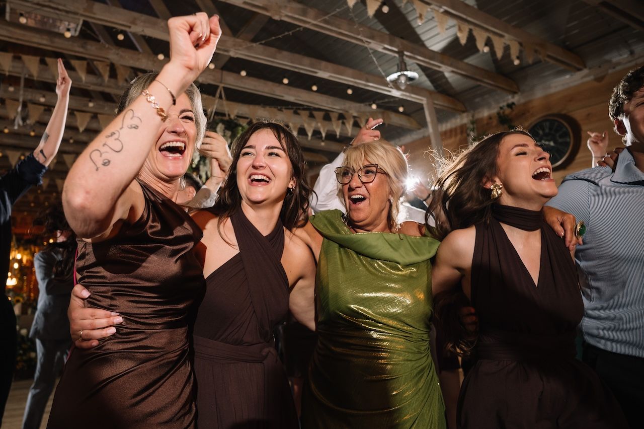 Four women in elegant dresses cheering and laughing together on a wedding dance floor.