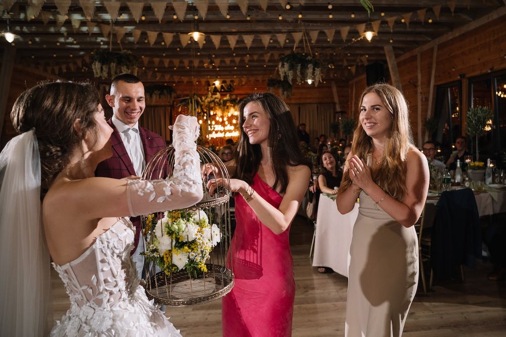 Bride holding floral birdcage bouquet with groom and guests celebrating at elegant rustic wedding reception.