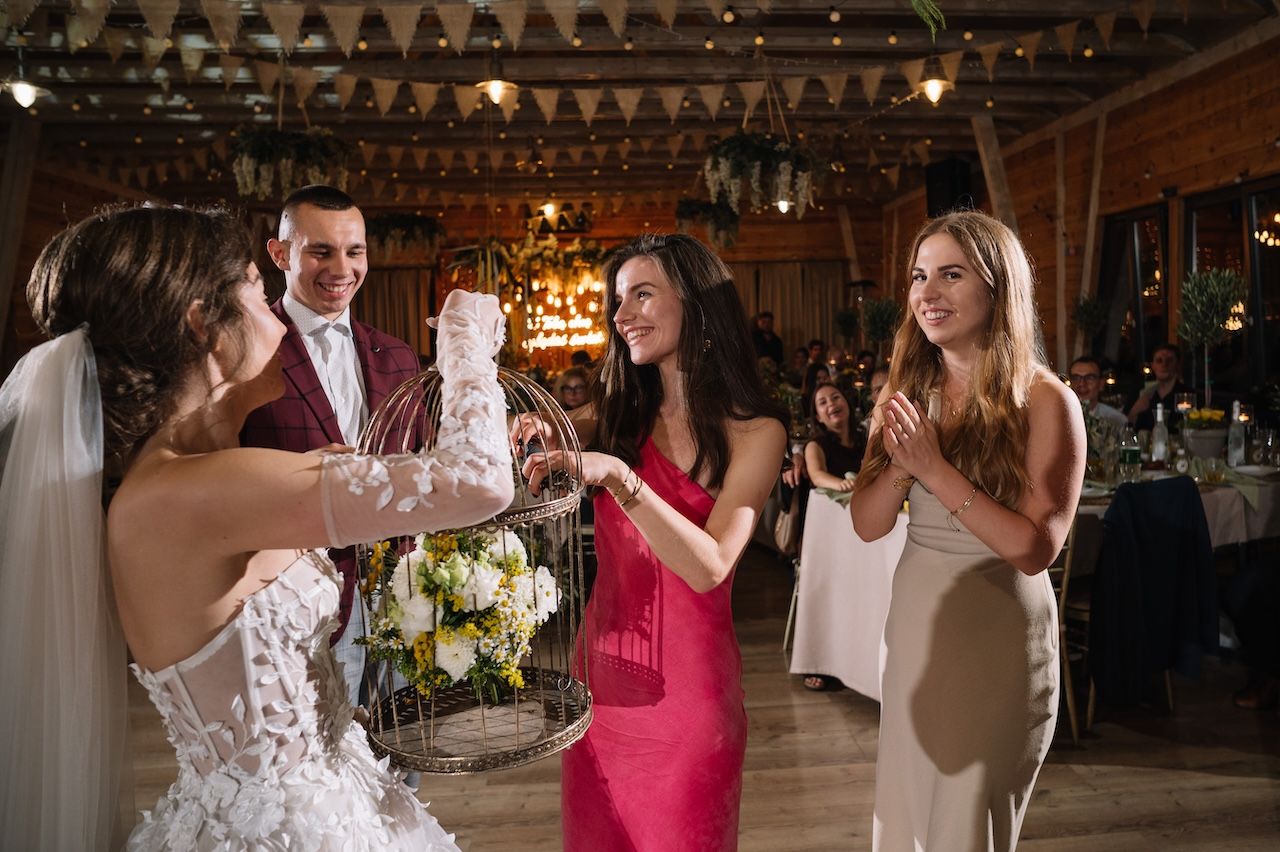 Bride holding floral birdcage bouquet with groom and guests celebrating at elegant rustic wedding reception.