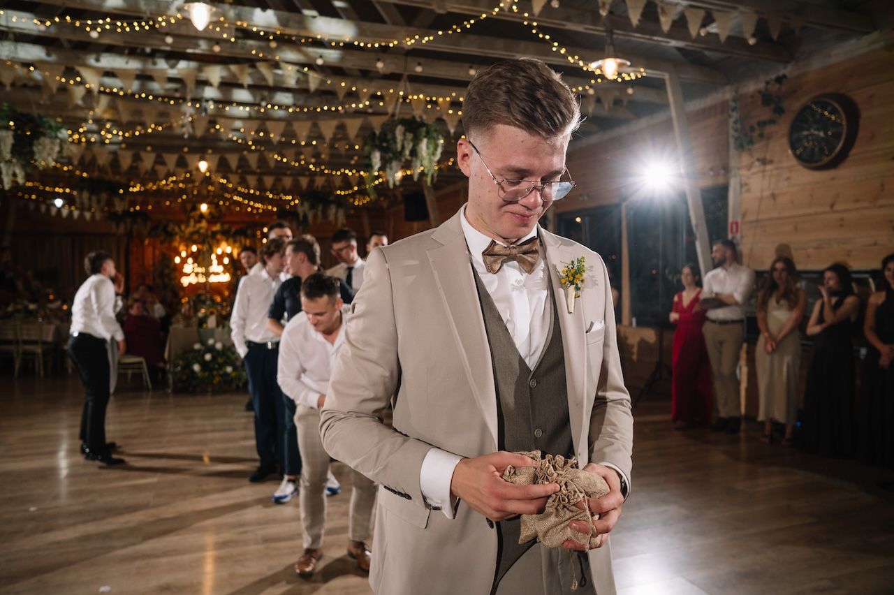 Groom in tan suit holding garter at rustic barn wedding reception with fairy lights.