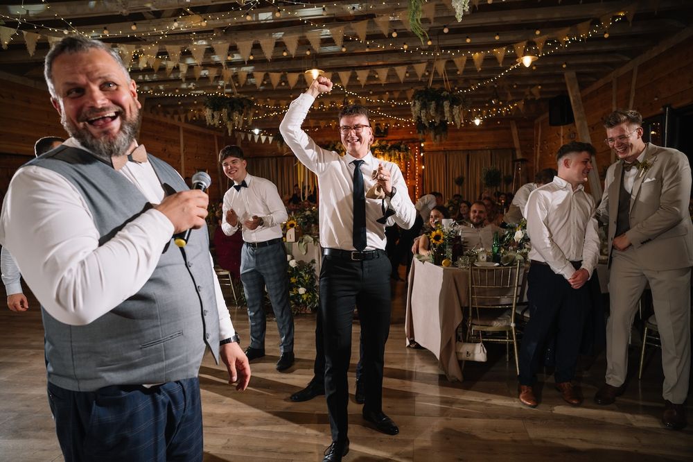 Men celebrating at wedding reception in rustic barn venue with string lights and decor.