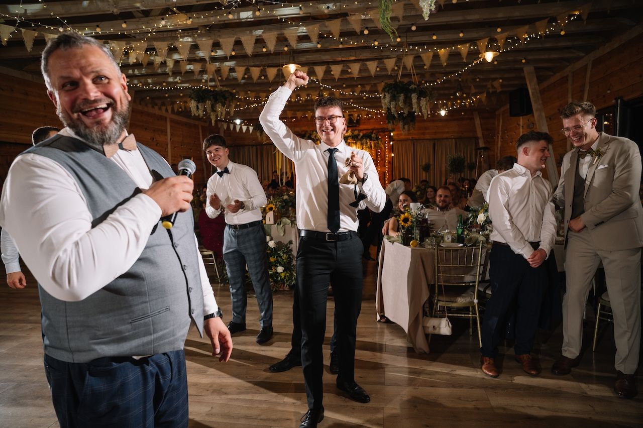 Men celebrating at wedding reception in rustic barn venue with string lights and decor.