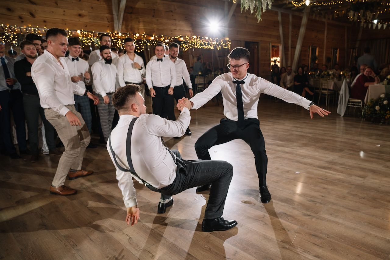 Groomsmen dancing energetically on wooden dance floor at rustic barn wedding reception.
