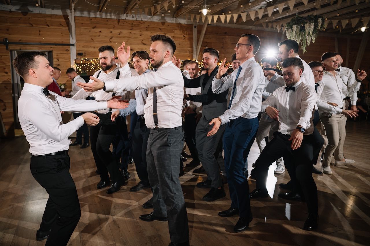 Large group of men dancing together on wooden floor at lively rustic barn wedding.