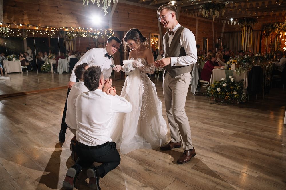 Groom kneeling before bride and groom during fun wedding reception tradition on dance floor.