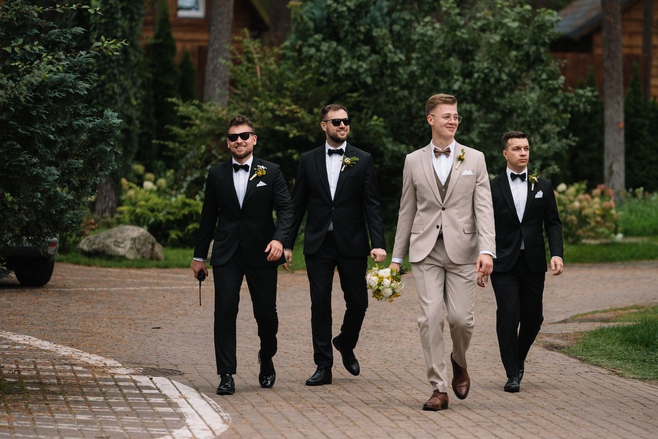 Four groomsmen walking together outdoors, groom in beige suit with three in black tuxedos.