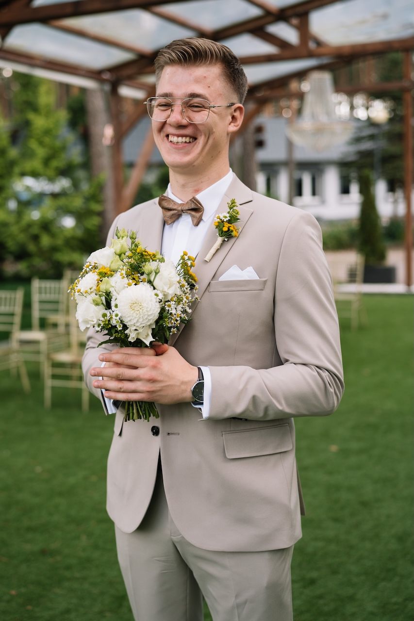 Happy groom in beige suit and bow tie holds white floral bouquet at outdoor wedding.