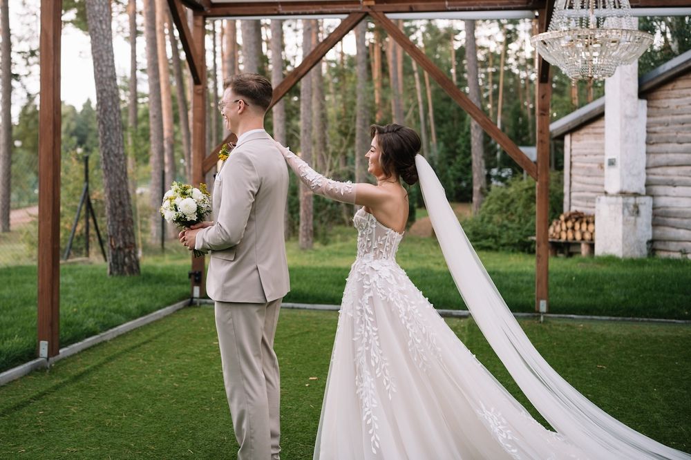 Bride in lace gown with long veil touches groom's shoulder during outdoor first look.