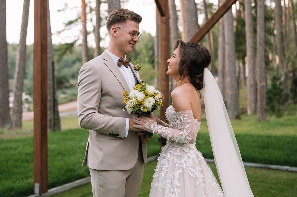 Smiling groom in beige suit holds bouquet while bride in lace dress laughs outdoors.
