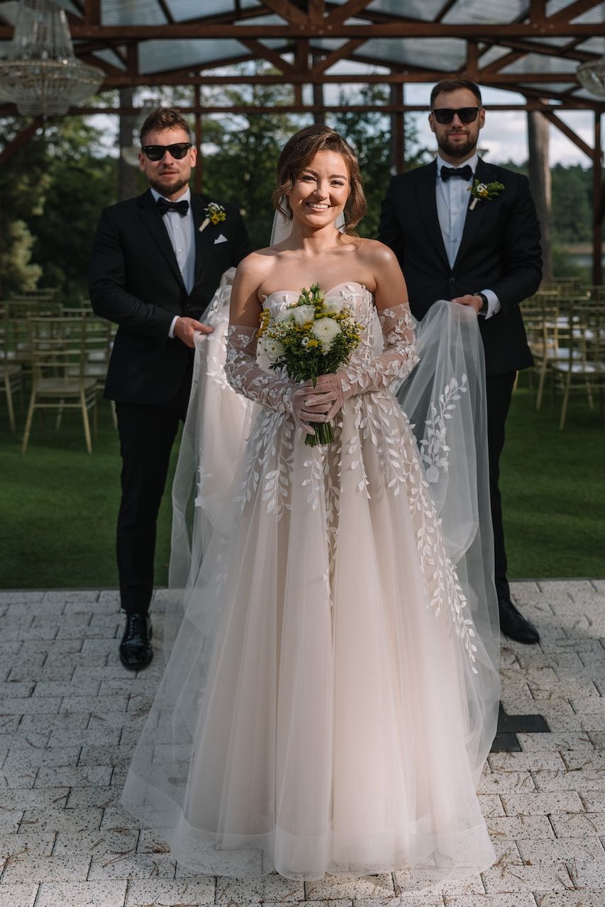 Smiling bride in floral lace wedding gown holds bouquet while two groomsmen in tuxedos hold her veil outdoors.