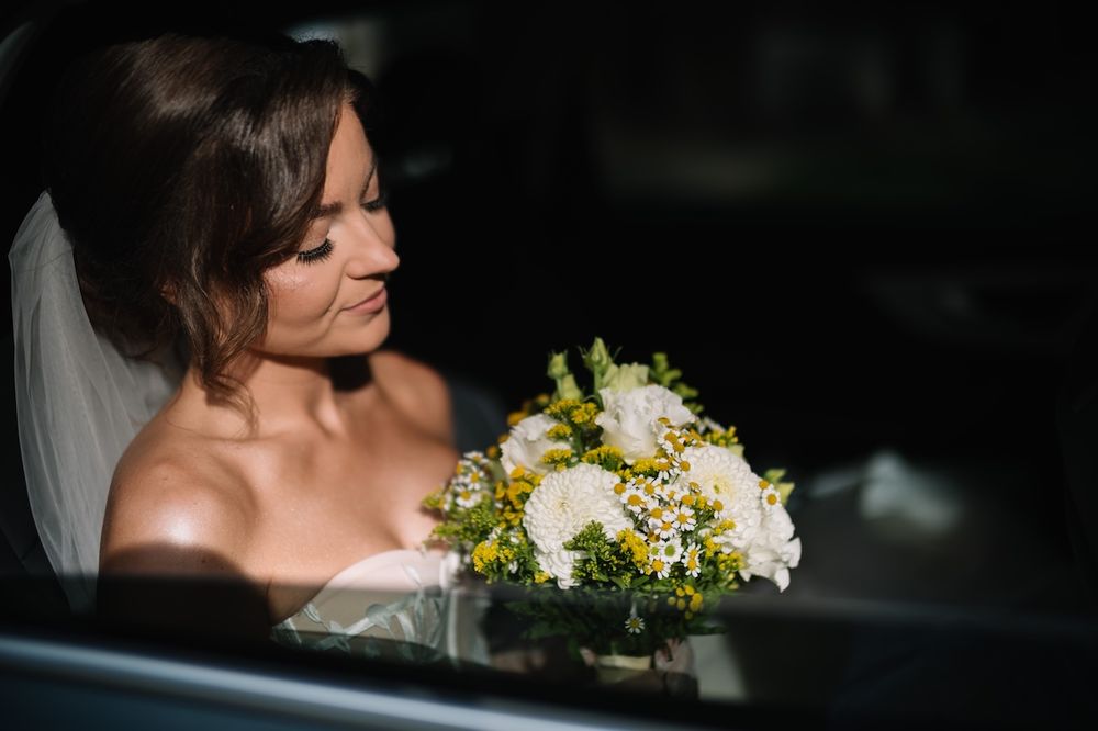 Elegant bride in white wedding dress holds yellow and white bouquet while seated in a car.