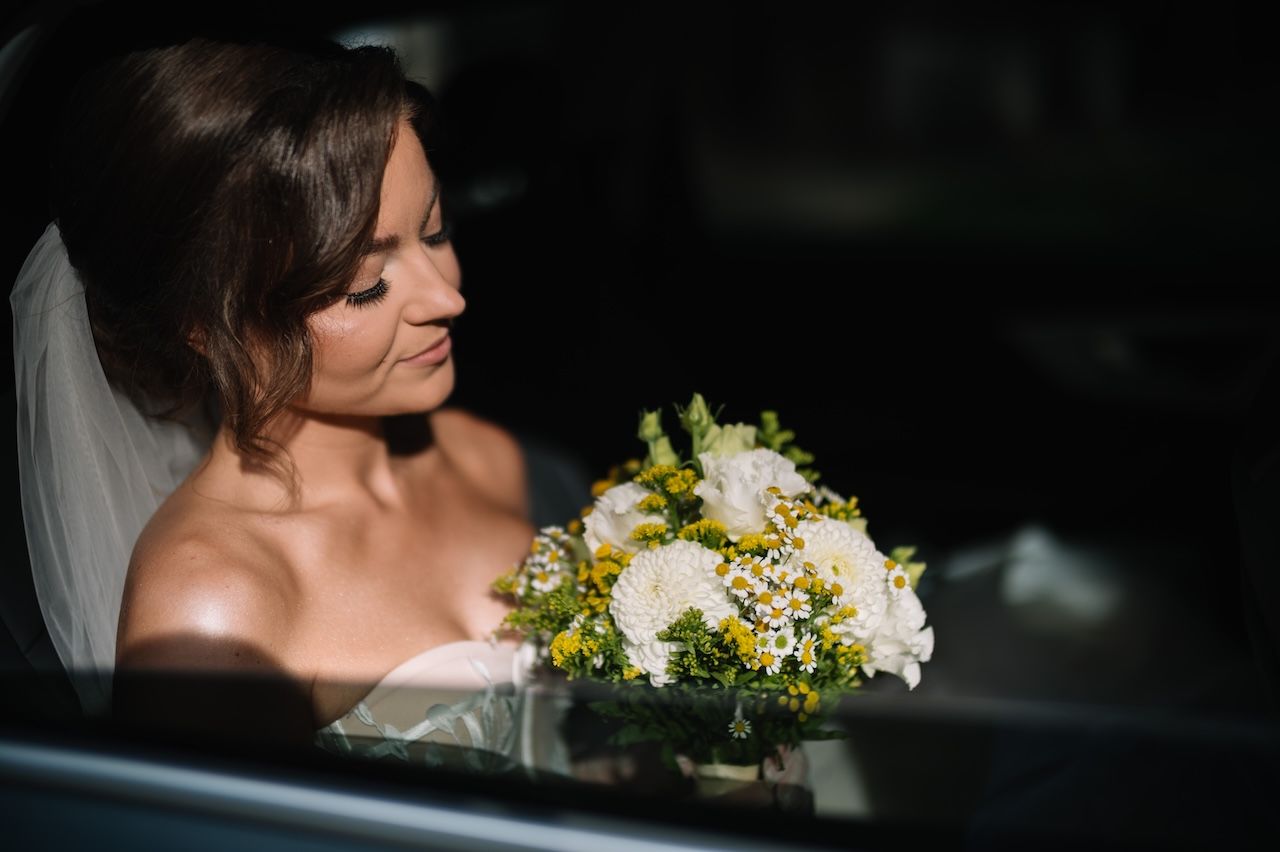 Elegant bride in white wedding dress holds yellow and white bouquet while seated in a car.