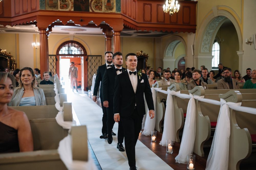 Groom in black tuxedo walks down candlelit church aisle with groomsmen and seated wedding guests.