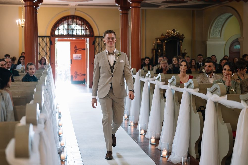 Groom in beige suit and bow tie walks confidently down a candlelit church aisle.