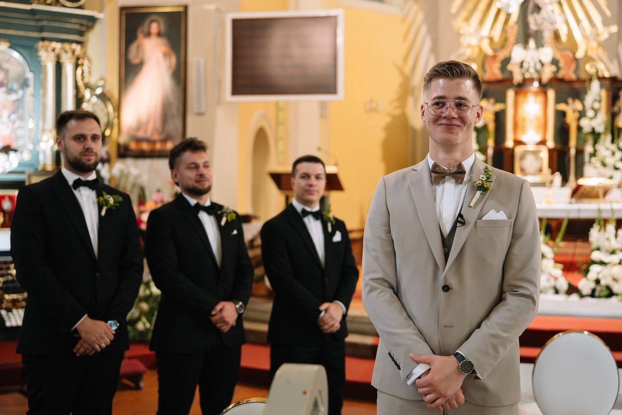 Groom in beige suit stands at church altar with three groomsmen in black tuxedos behind him.