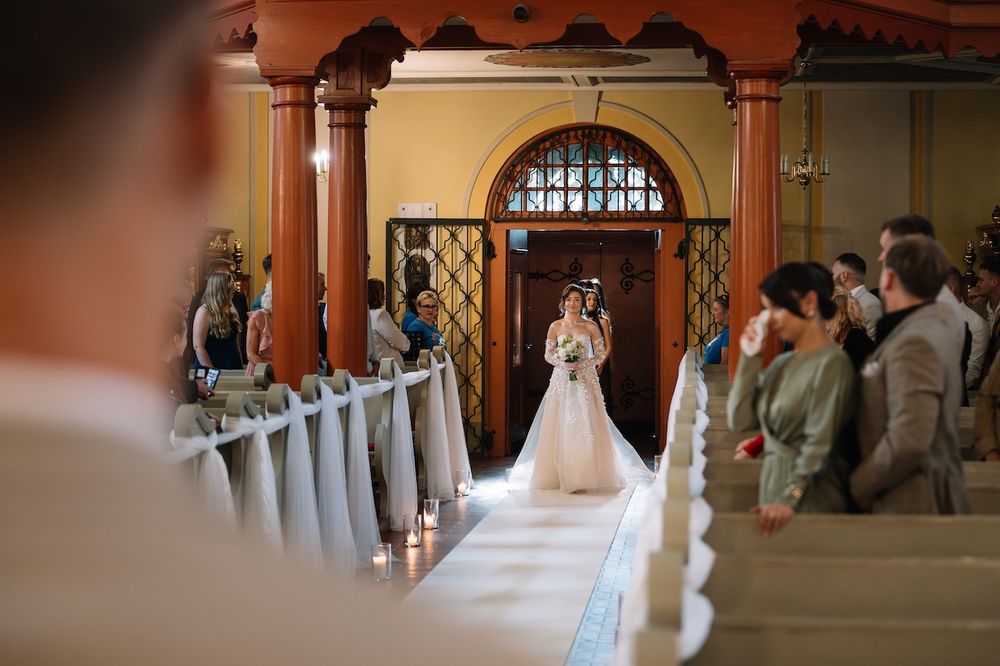Bride in white gown walks down candlelit church aisle toward waiting groom and guests.