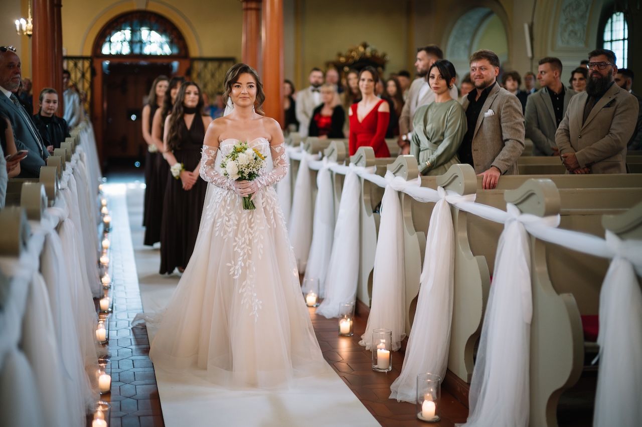 Bride walking down decorated church aisle holding bouquet with guests watching.