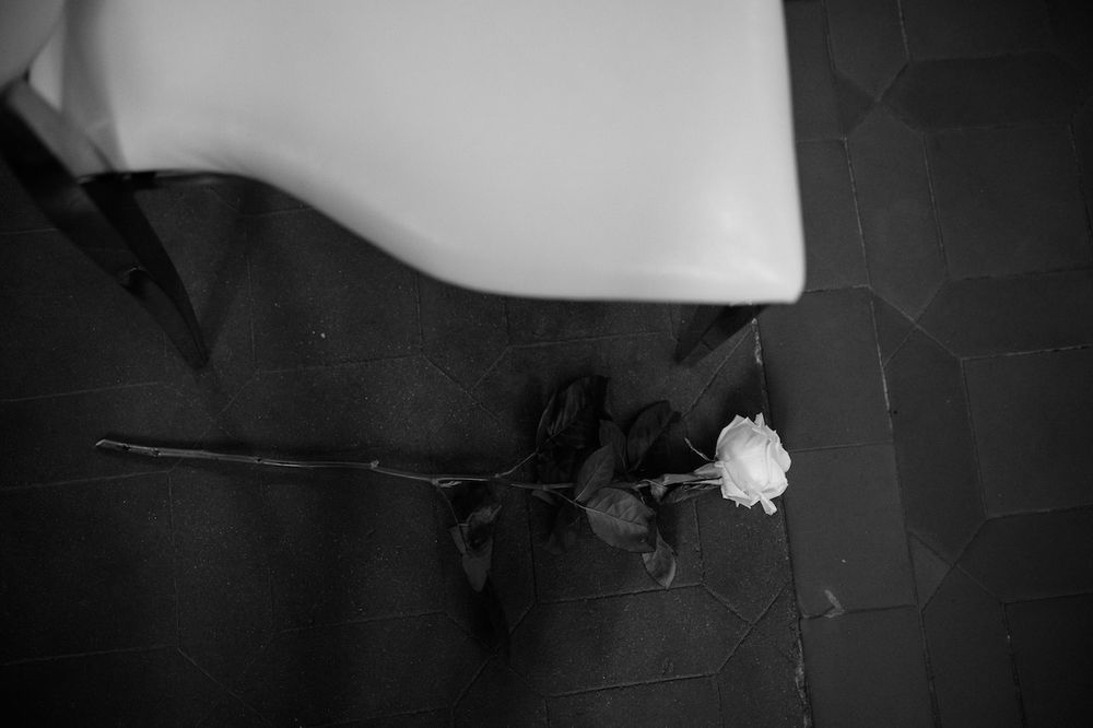 Single white rose lying on dark tile floor beside white furniture in black and white.