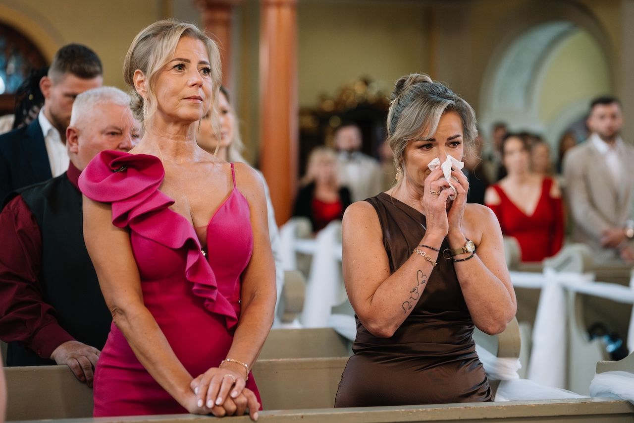 Two emotional female wedding guests crying during church ceremony, one in pink dress.
