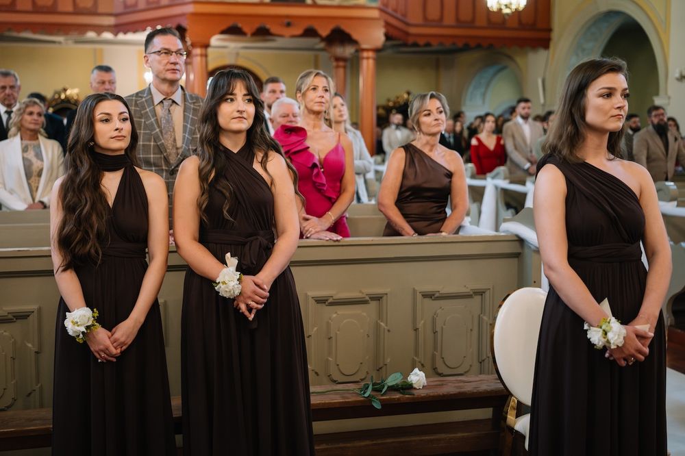 Three bridesmaids in black dresses standing in church during wedding ceremony.