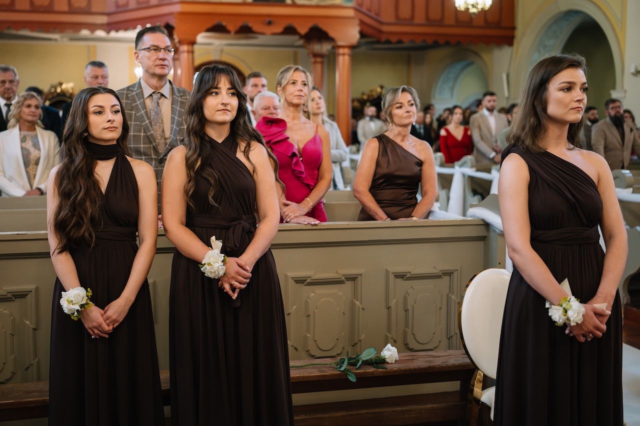 Three bridesmaids in black dresses standing in church during wedding ceremony.