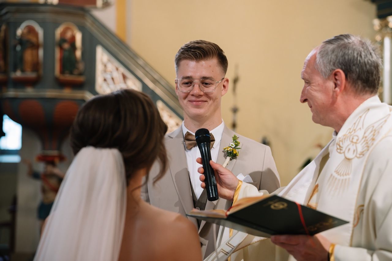 Groom smiling while reciting wedding vows to bride with priest holding book.