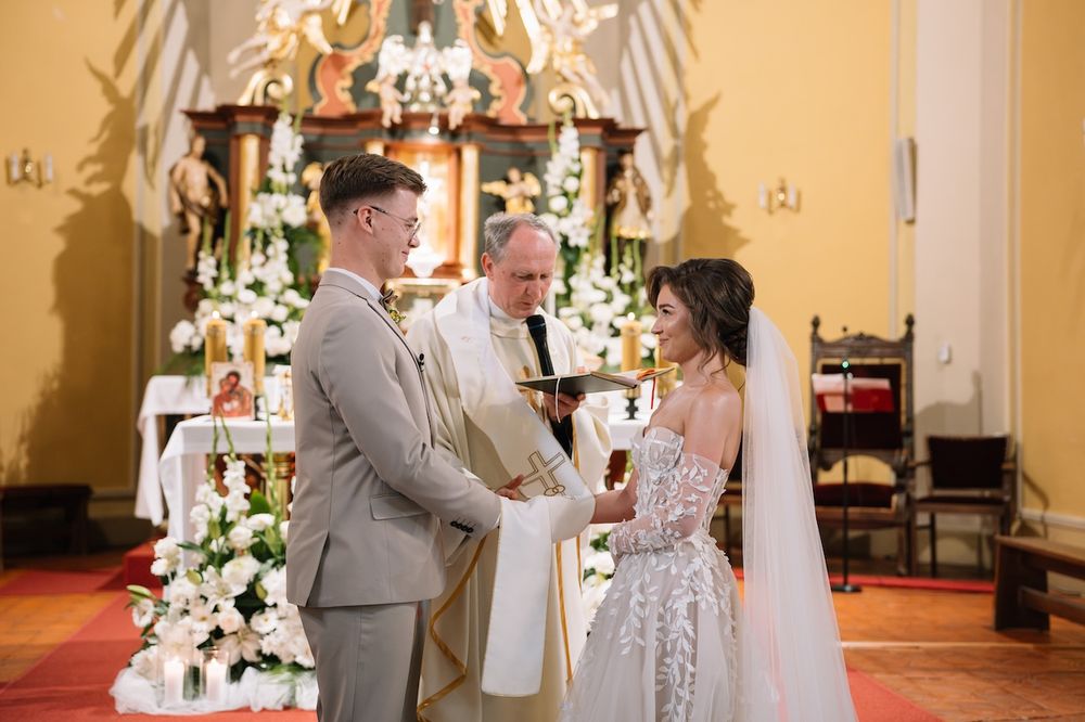 Bride and groom exchanging vows before priest at elegant church altar adorned with white flowers.
