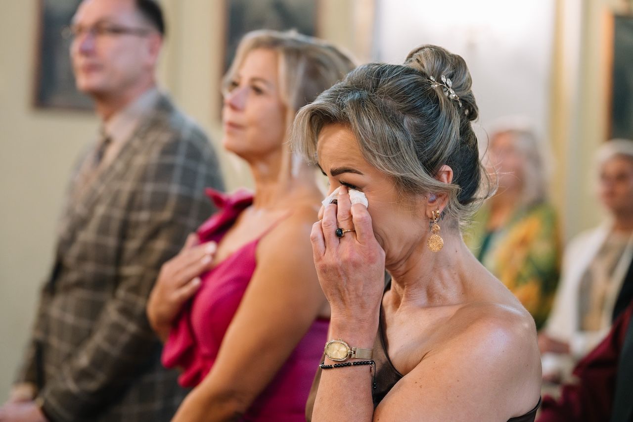 Emotional wedding guest wiping tears during church ceremony while others stand in background.