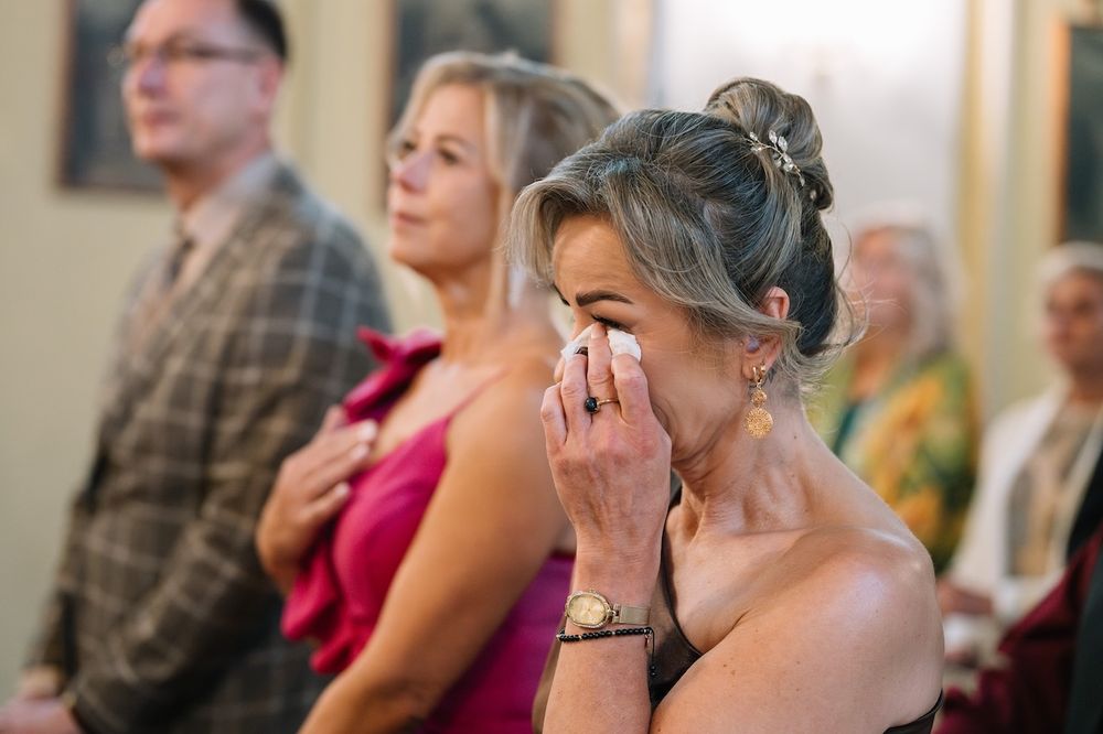 Emotional wedding guest wiping tears during church ceremony while others stand in background.