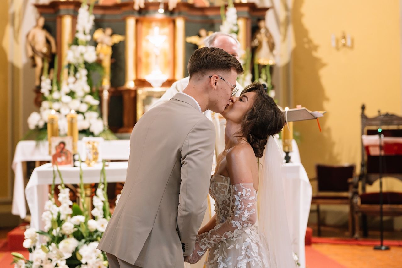 Bride and groom sharing first kiss at church altar surrounded by white floral arrangements.
