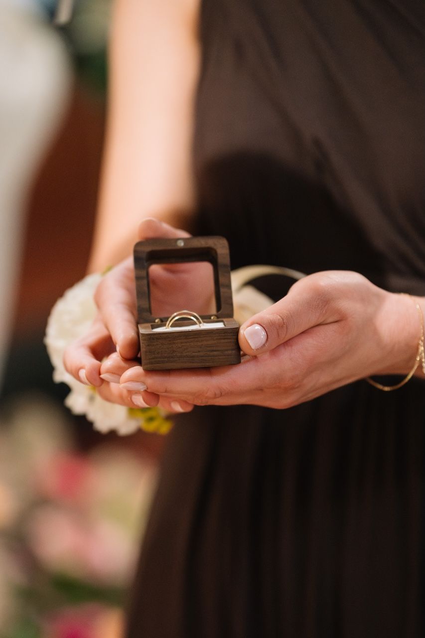 Bridesmaid in black dress holding wooden ring box with wedding bands during ceremony.