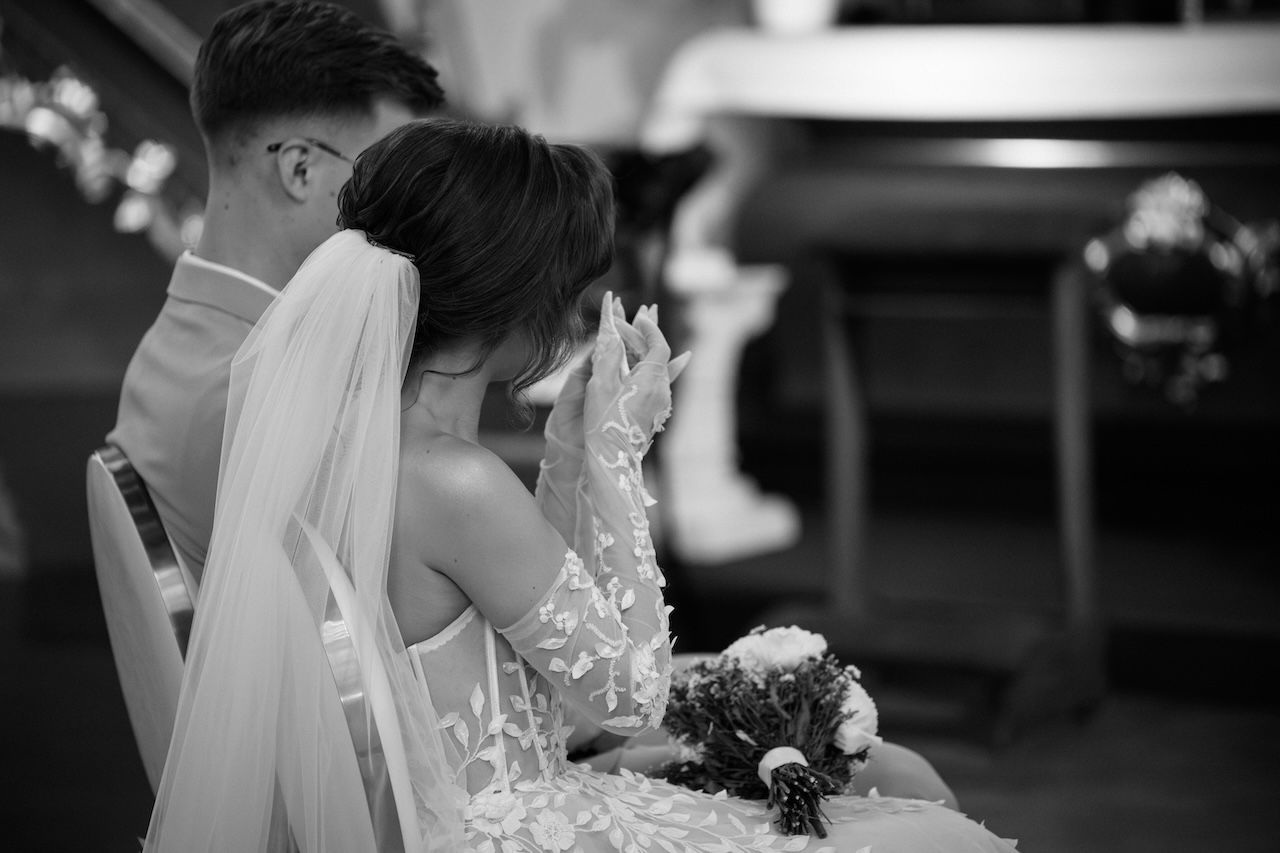Black and white photo of bride wiping tears while holding bouquet beside groom in church.