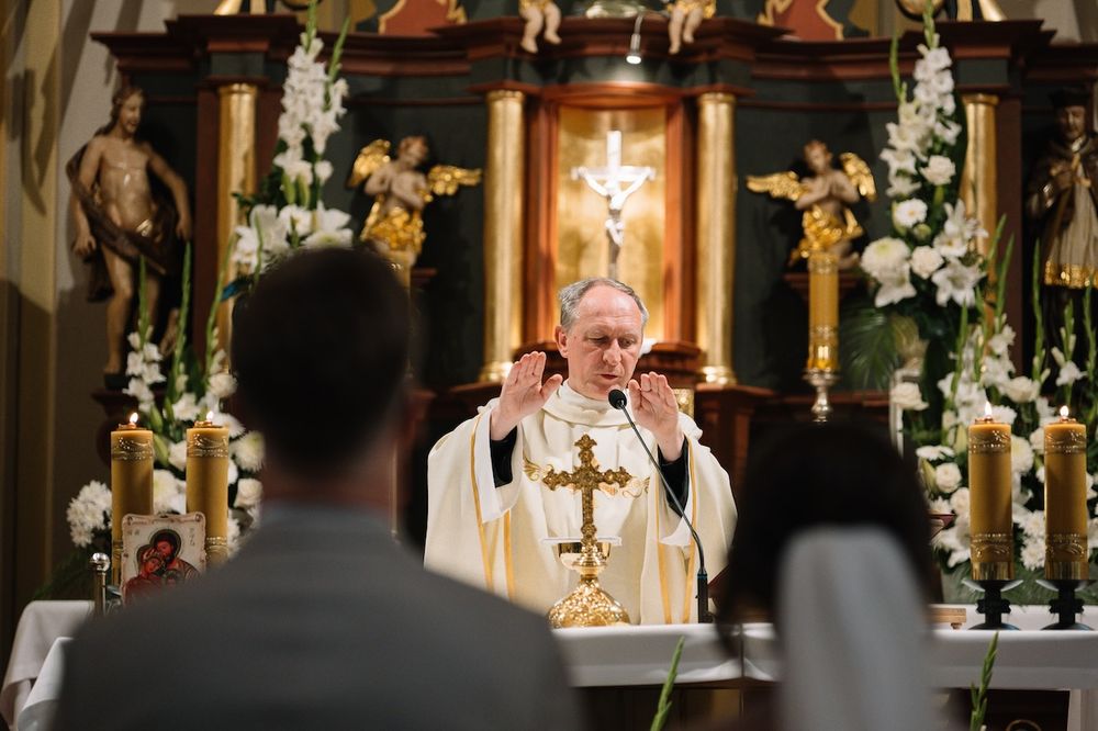 Catholic priest raising hands while delivering wedding sermon at ornate church altar with cross.