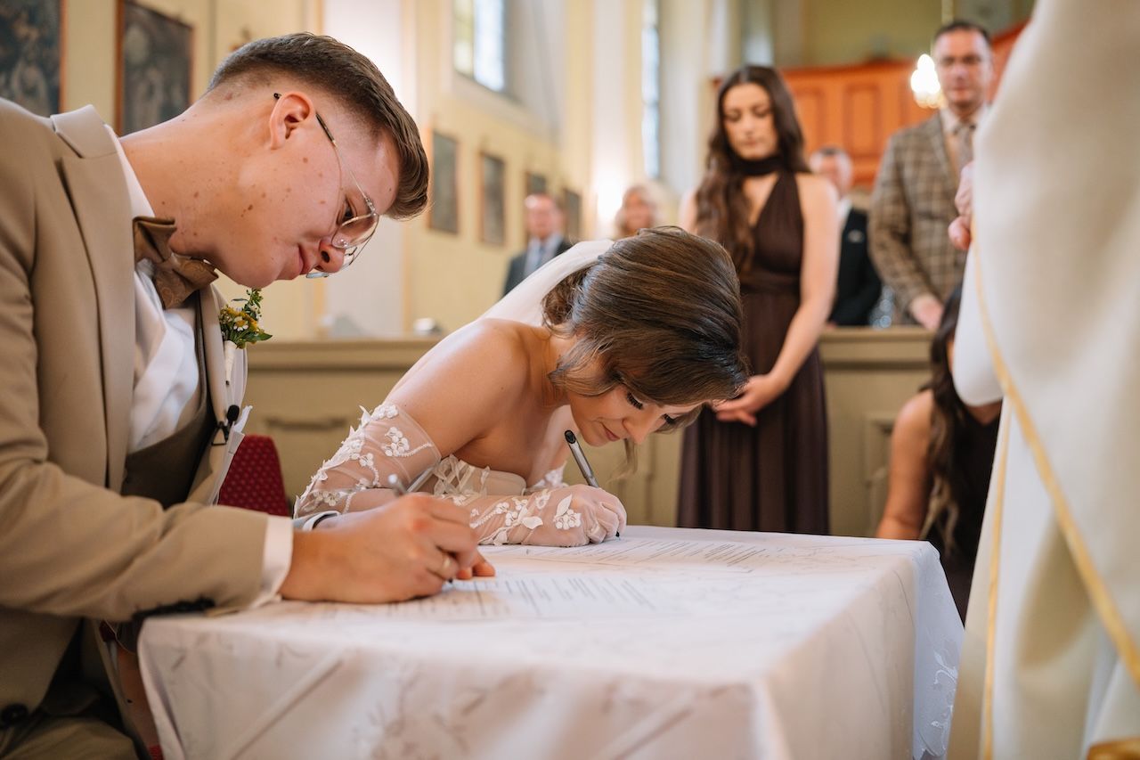 Bride signing wedding registry documents at church altar with groom beside her during ceremony.