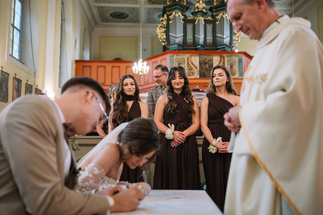 Bride and groom signing marriage certificate at church with bridesmaids and priest watching.