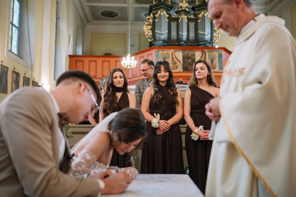 Bride and groom signing marriage certificate at church with bridesmaids and priest watching.