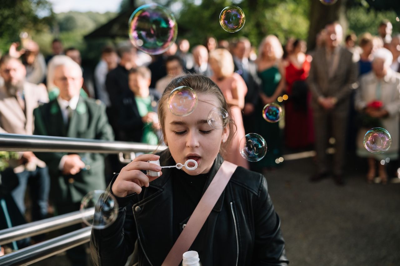 Young girl blowing colorful soap bubbles at outdoor wedding celebration with guests behind her.