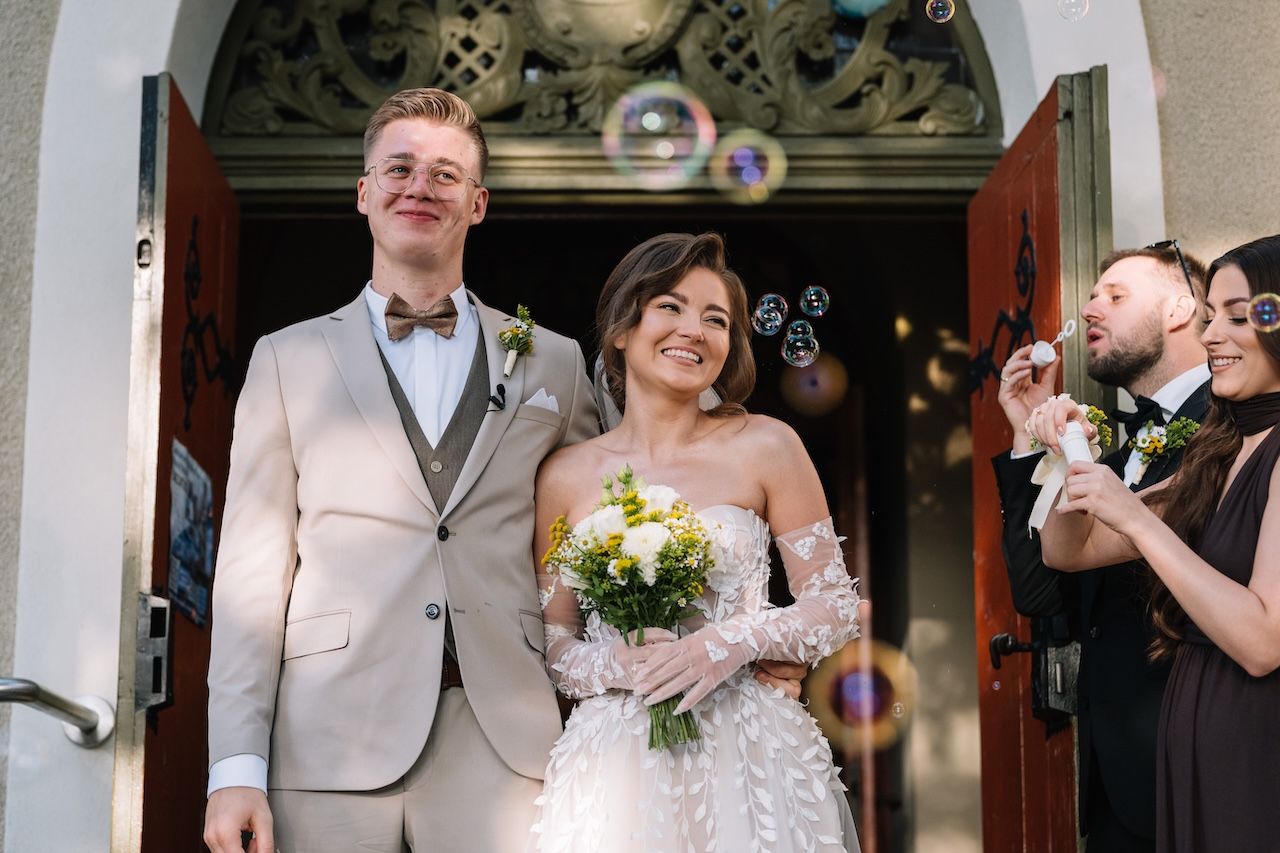 Smiling newlyweds exit church doorway surrounded by soap bubbles and cheering wedding guests.