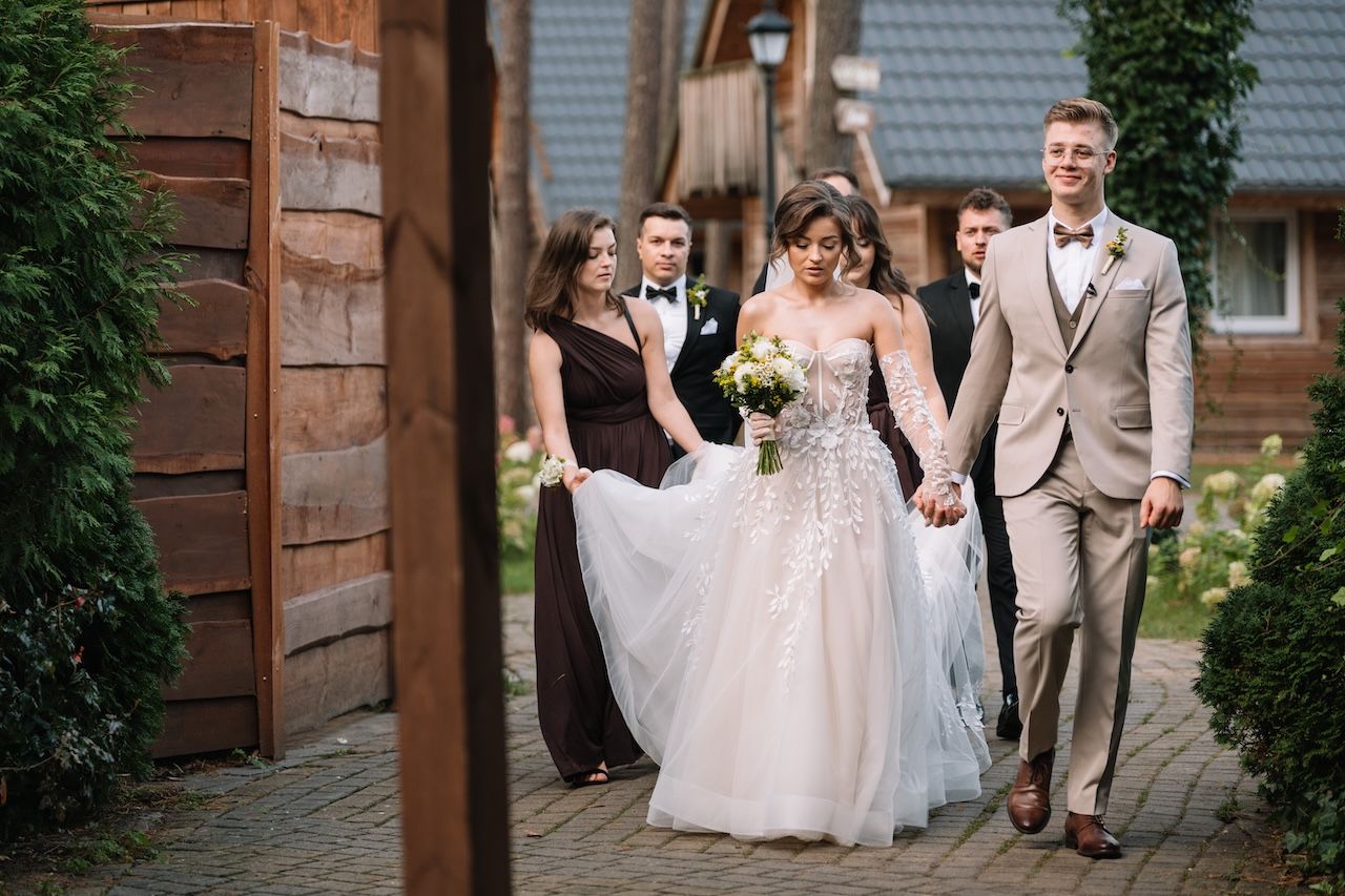 Bride and groom walk hand in hand with wedding party along rustic cobblestone path.