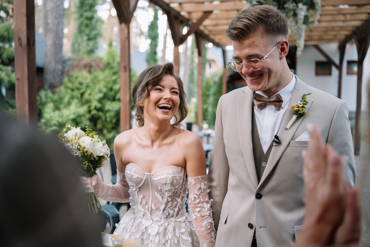 Joyful bride laughing with groom at outdoor wedding reception surrounded by celebrating guests.