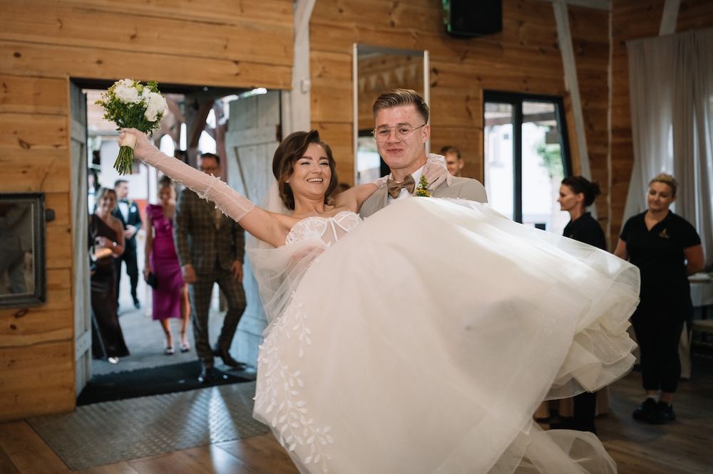 Groom carries bride in white wedding dress through venue entrance as guests celebrate their arrival.